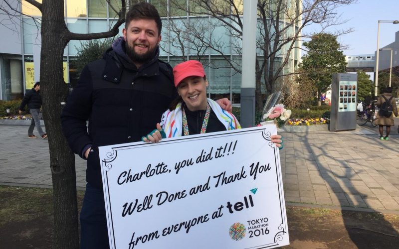 A photo of Charlotte Shanks holding a board of "Charlotte, you did it!! Well Done and Thank you from everyone at Tell" and a man standing besides her