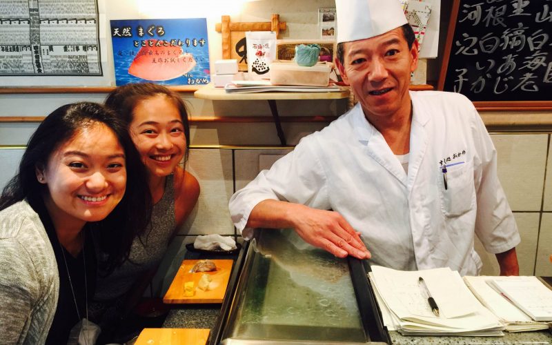 Two women smiling at a sushi chef across the counter